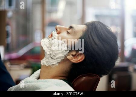 Profil du client masculin assis au salon de coiffure avec rasage crème sur le visage Banque D'Images