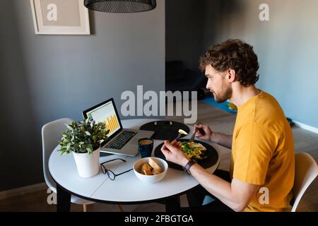 Homme indépendant ayant de la nourriture tout en travaillant sur un ordinateur portable à la maison Banque D'Images
