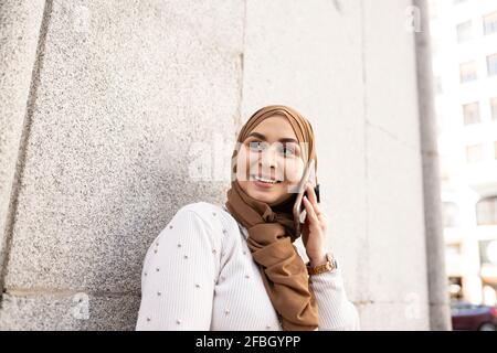 Jeune femme dans le hijab regardant loin tout en parlant sur intelligent téléphone au mur Banque D'Images