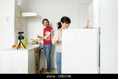 Femme souriante ouvrant la porte du réfrigérateur pendant que l'homme prépare des aliments cuisine à la maison Banque D'Images