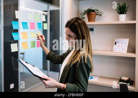 Femme d'affaires de taille moyenne souriante tenant une planchette à pince tout en planifiant la stratégie note adhésive au bureau Banque D'Images