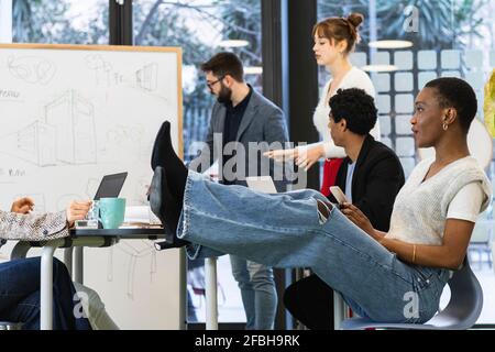 Femme d'affaires avec téléphone mobile, les jambes sont penchées sur la table avec des collègues en arrière-plan au bureau Banque D'Images