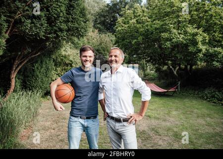 Fils souriants avec un ballon de basket debout avec un père dans l'arrière-cour Banque D'Images