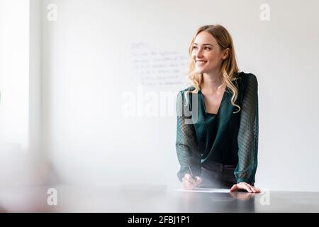 Femme d'affaires souriante regardant loin tout en écrivant sur un document lors d'une conférence tableau Banque D'Images