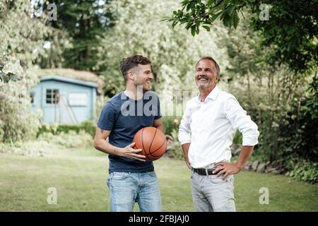 Père souriant avec les mains sur la hanche debout avec le fils tenant basket-ball dans l'arrière-cour Banque D'Images