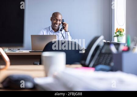 Homme d'affaires souriant parlant sur un téléphone fixe tout en étant assis au bureau au bureau Banque D'Images
