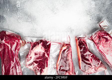 Viande de bœuf crue dans l'ensemble de peaux aspirées, tomahawk, t-bone, steak de club, coupes d'oeillet et de filet de côtes, sur fond de pierre grise, plan à plat avec vue de dessus Banque D'Images