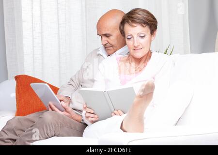 Couple marié d'âge moyen se détendant à la maison lisant sur un canapé avec l'homme utilisant une tablette-pc comme la femme aime un livre dans un portrait de haute clé Banque D'Images