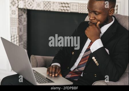 Beau homme d'affaires afro-américain en costume classique utilise un ordinateur portable et souriant dans un bureau moderne Banque D'Images