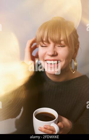 Jolie femme blonde caucasienne portant un chandail noir et de belles boucles d'oreilles tenant une tasse de café et rire. Effet de lumière floue. Ambiance romantique ou concept café du matin. Photo de haute qualité Banque D'Images