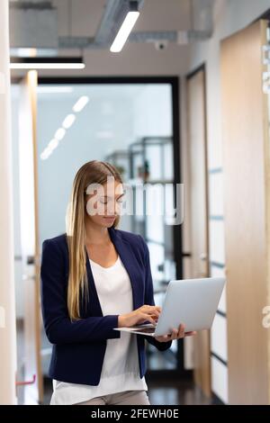 Femme d'affaires caucasienne élégamment habillée debout dans un couloir à l'aide d'un ordinateur portable Banque D'Images