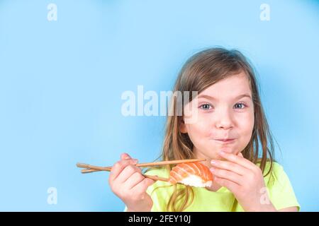 Une petite fille adorable aime manger des sushis. Joyeux drôle caucasien blonde petite fille avec divers rouleaux de sushi et baguettes. Sur fond bleu vif Banque D'Images
