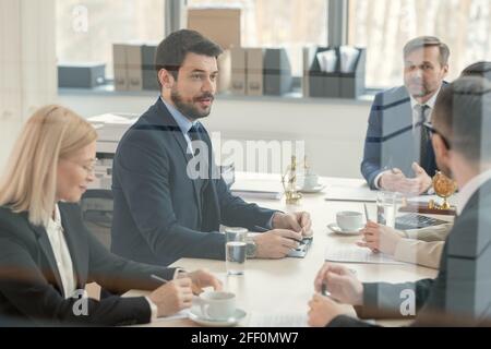 Jeune avocat confiant en costume assis à la table de conférence et discutant du contenu du contrat avec ses collègues, derrière une vue en verre Banque D'Images