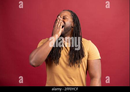 Un gars afro-américain endormi bâtit et se sent ennuyé isolé sur fond rouge, l'homme noir fatigué ressent un manque d'énergie et de sommeil, un homme fatigué a besoin de se reposer Banque D'Images