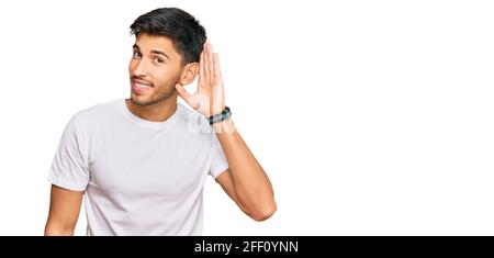 Jeune homme beau portant un tshirt blanc décontracté souriant de la main sur l'oreille écoutant une audition à la rumeur ou des potins. Concept de surdité. Banque D'Images