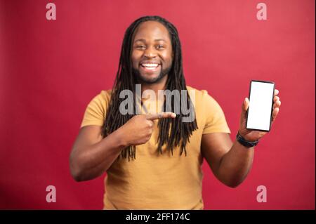 Homme afro-américain avec des dreadlocks se tenant isolé sur le fond rouge, présentant une nouvelle application, pointant avec les mains sur le téléphone mobile avec un espace blanc pour la publicité, image maquette Banque D'Images