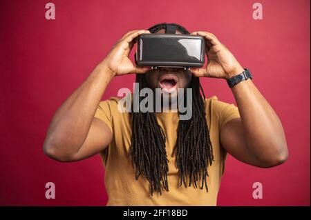Homme noir émerveillé avec de longues dreadlocks vêtues de vêtements décontractés, debout isolé sur fond rouge avec casque de réalité virtuelle, lunettes dans une pièce vide, voyager en ligne, essayer une nouvelle expérience Banque D'Images