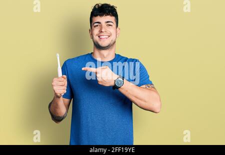 Jeune homme hispanique tenant une brosse à dents électrique souriant avec qui il se fait plaisir de pointer main et doigt Banque D'Images