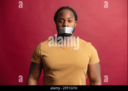 Le concept de racisme avec un homme africain en colère avec une bouche scellée avec la bande regardant la caméra, isolé sur fond rouge, sans voix, a énervé Guy en raison de l'incapacité de dépasser son opinion Banque D'Images