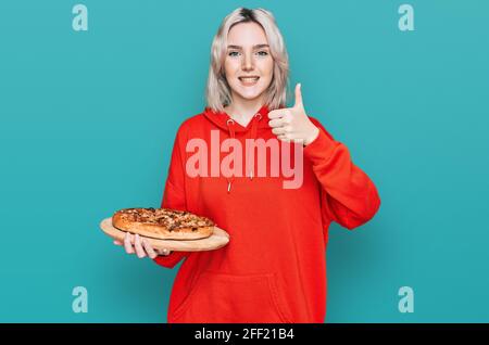 Jeune fille blonde tenant une pizza italienne souriante heureuse et positive, pouce vers le haut faisant excellent et signe d'approbation Banque D'Images