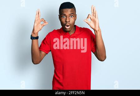 Jeune afro-américain portant un t-shirt rouge décontracté à l'aspect surpris et choqué faisant ok symbole d'approbation avec les doigts. Expression folle Banque D'Images