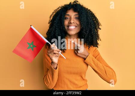 Femme afro-américaine aux cheveux afro tenant le drapeau marocain souriant contenchez-vous de pointer avec votre main et votre doigt Banque D'Images