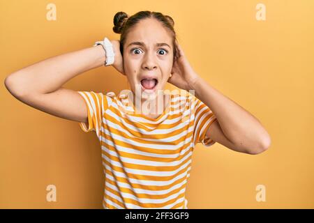 Belle brune petite fille portant décontracté t-shirt rayé fou et peur avec les mains sur la tête, peur et surpris de choc avec la bouche ouverte Banque D'Images