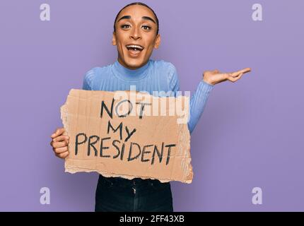 Homme transgenre hispanique portant un maquillage et de longues cheveux tenue ce n'est pas la bannière de protestation de mon président qui célèbre la victoire avec un sourire heureux et le gagnant express Banque D'Images