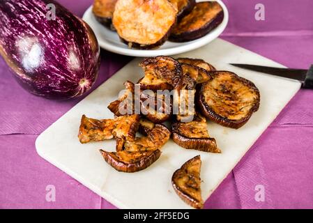 Aubergine frite tranchée sur le plan de cuisine avec couteau, cercles d'aubergine cuits sur petite assiette blanche et aubergine crue, le tout sur fond violet. Banque D'Images