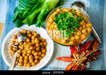 Pois chiches bouillis sur l'assiette et dans un bol en verre, bouquet de piments rouges séchés, cuillère sur table bleue, vue du dessus. Banque D'Images