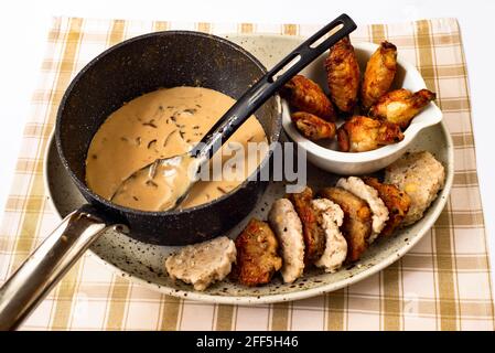 Sauce aux champignons dans une casserole avec une cuillère, cuisses de poulet grillées dans un petit bol, boulettes (cuites et frites en alternance) sur une assiette sur la table. Banque D'Images