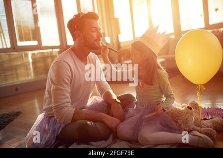 Une petite fille met le maquillage sur le visage de son père pendant qu'ils se préparent à des exercices de ballet dans une atmosphère détendue à la maison. Famille, ensemble, maison Banque D'Images