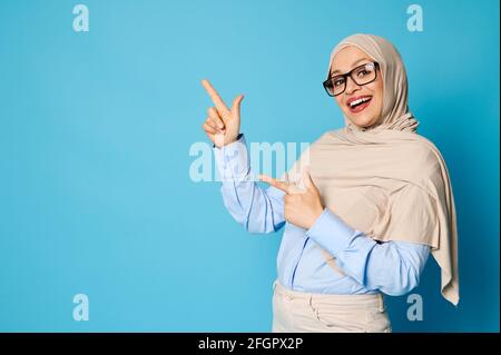 Charmante femme arabe dans hijab avec beau sourire la pointe index sur fond bleu avec espace de copie Banque D'Images