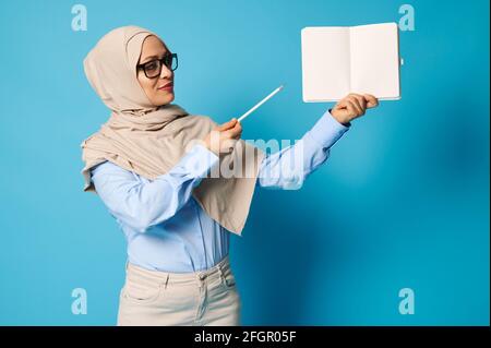 La femme musulmane dans le hijab tient un livre blanc et des points à avec un stylo isolé sur fond bleu avec espace de copie Banque D'Images