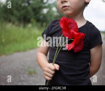 Un jeune garçon triste dans un T-shirt noir tient deux fleurs de pavot dans ses mains. Concentrez-vous sur la fleur. Réflexion sur le sujet de la guerre. Memorial Day Banque D'Images