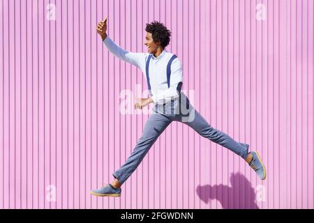 Homme noir, avec des cheveux afro, prenant un selfie avec son smartphone tout en sautant sur un fond de volets roses. Banque D'Images