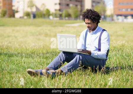 Homme de travail noir aux cheveux d'afro à l'aide de son ordinateur portable assis sur un skateboard sur l'herbe d'un parc urbain. Banque D'Images