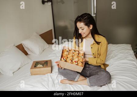 Charmante femme assise sur le lit avec des boîtes de nourriture pleines de croissant, a un dîner romantique à la maison Banque D'Images