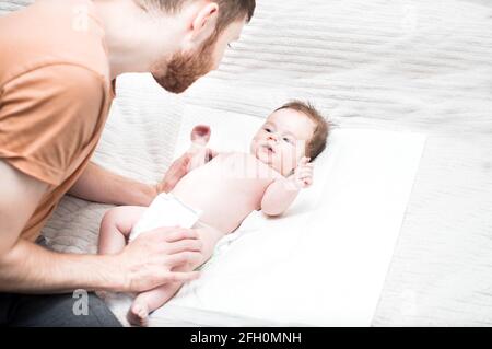 le jeune père moderne s'occupe de son fils nouveau-né. Change la couche sur le lit. Concept d'hygiène et de soins aux nouveau-nés. L'amour et le soin du père. Le père d Banque D'Images