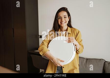 Une jeune femme heureuse sourit et tient un robot-aspirateur à la maison Banque D'Images