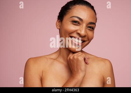 Portrait d'une femme à peau brune souriant sur fond rose. Belle femme avec une peau saine regardant l'appareil photo avec la main sur le menton. Banque D'Images