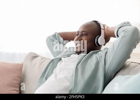 Afro-américain d'âge moyen dans un casque sans fil écoutant de la musique sur un canapé dans le salon, espace de copie Banque D'Images