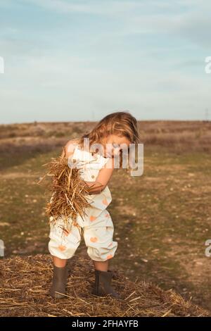 Adorable enfant en combinaison jouant avec du foin près des balles de paille en campagne Banque D'Images