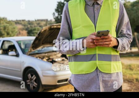 Utilisez un gilet de sécurité vert pour la récolte d'un conducteur mâle méconnaissable téléphoner et demander de l'aide après un accident de voiture dans le pays route Banque D'Images