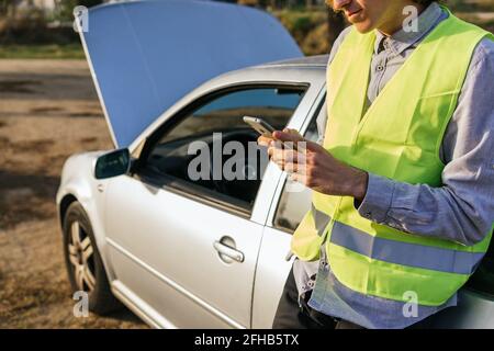 Utilisez un gilet de sécurité vert pour la récolte d'un conducteur mâle méconnaissable téléphoner et demander de l'aide après un accident de voiture dans le pays route Banque D'Images