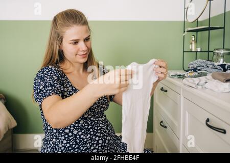 Femme joyeuse en robe décorative assise sur le ballon de yoga avec des vêtements pour enfant contre la mode dans la chambre Banque D'Images