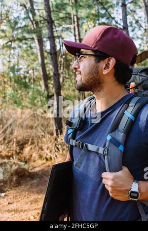Un routard mâle barbu dans le cap marchant parmi les arbres et les plantes dans les bois en journée ensoleillée Banque D'Images