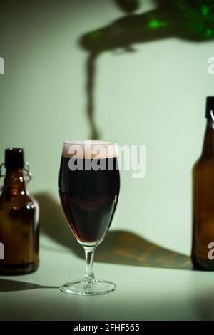 Bière sombre déversant d'une bouteille à un verre avec tête en mousse sur fond vert coloré, ombres vives des feuilles de palmier Banque D'Images