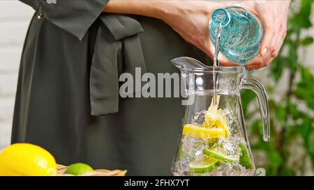 Buvez de l'eau. Main de femme versant de l'eau pure fraîche avec des morceaux de citron du pichet dans le verre dans le concept de santé et de régime du matin. Une vie saine Banque D'Images