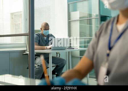 Un médecin bald occupé dans un masque chirurgical et des exfoliants assis à la table et en saisissant le rapport sur l'ordinateur portable en arrière-plan Banque D'Images